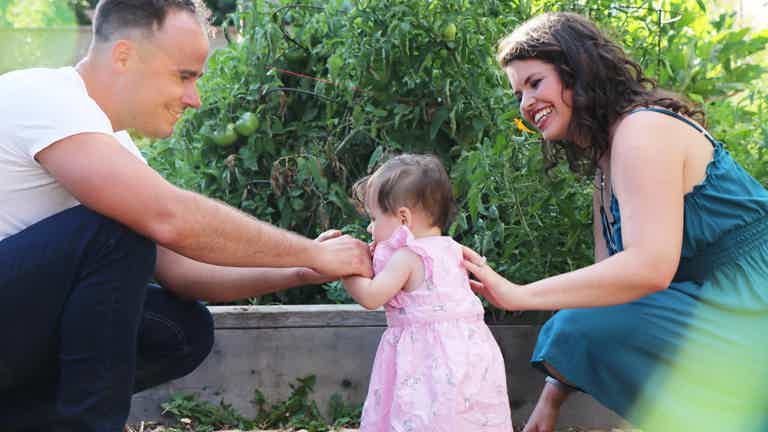 Parents and a toddler in garden, child learning to walk with support from both adults, smiles and encouragement.