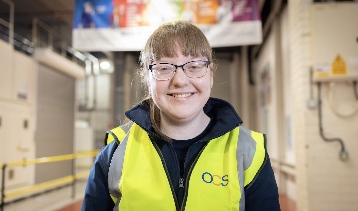 A person wearing glasses and a high-visibility vest with an OCS logo smiles while standing indoors in a well-lit industrial or warehouse setting.