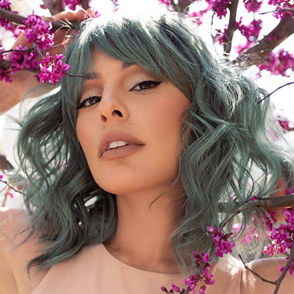  A woman with green haired wig in the style Breezy Wavez in shade Smoky Forest, is standing in front of a tree with purple flowers.