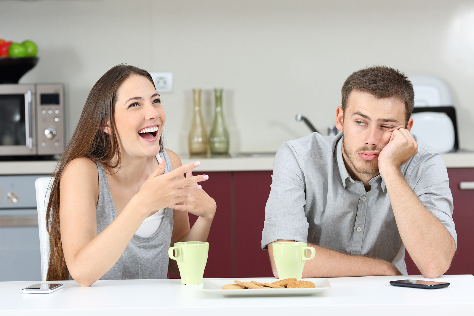 Photo of a young white man and woman wearing gray shirts and talking in a kitchen. She is laughing while he is looking annoyed at her.
