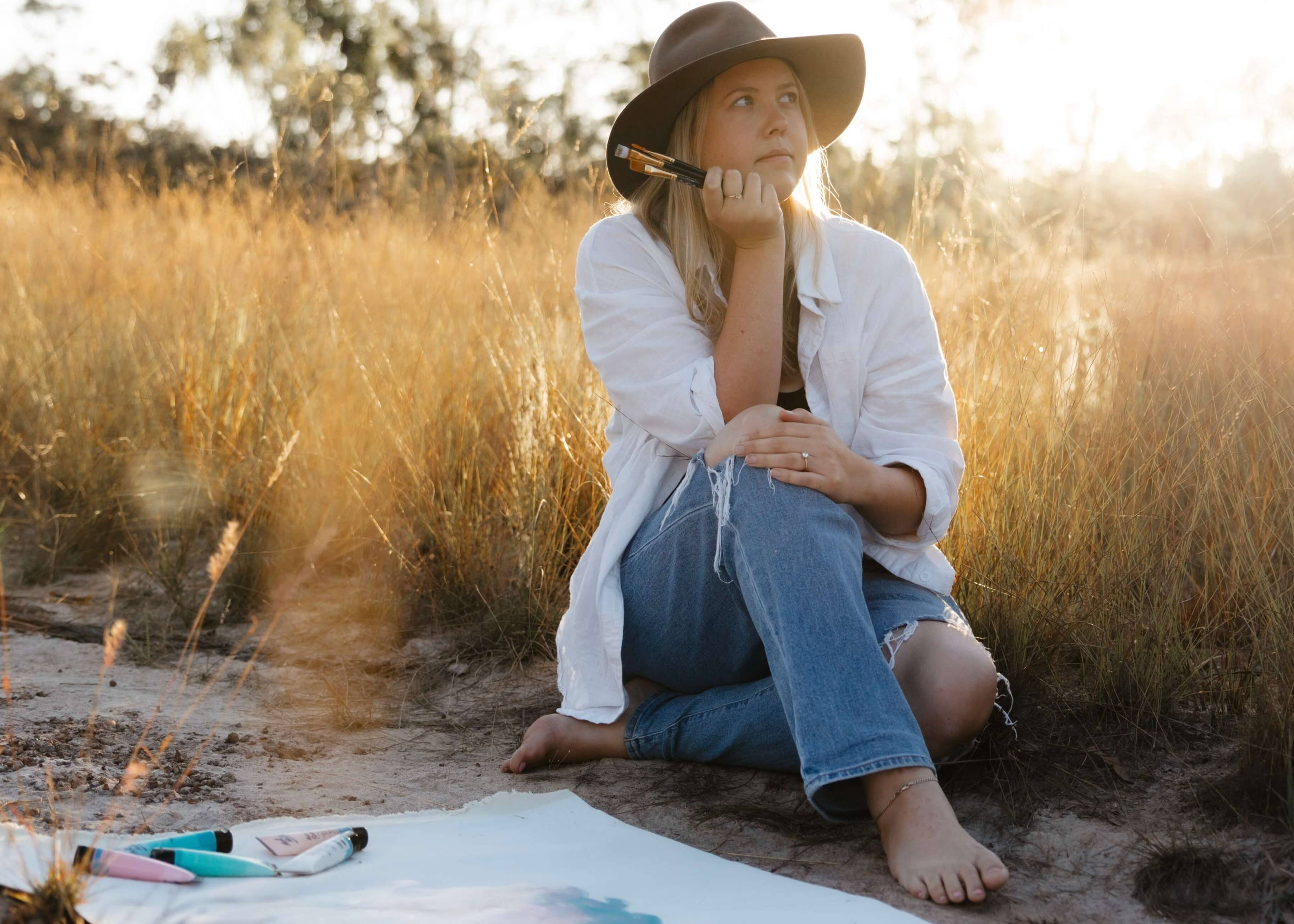 Caitlin Trindall sat in front of grass and a canvas holding paint brushes