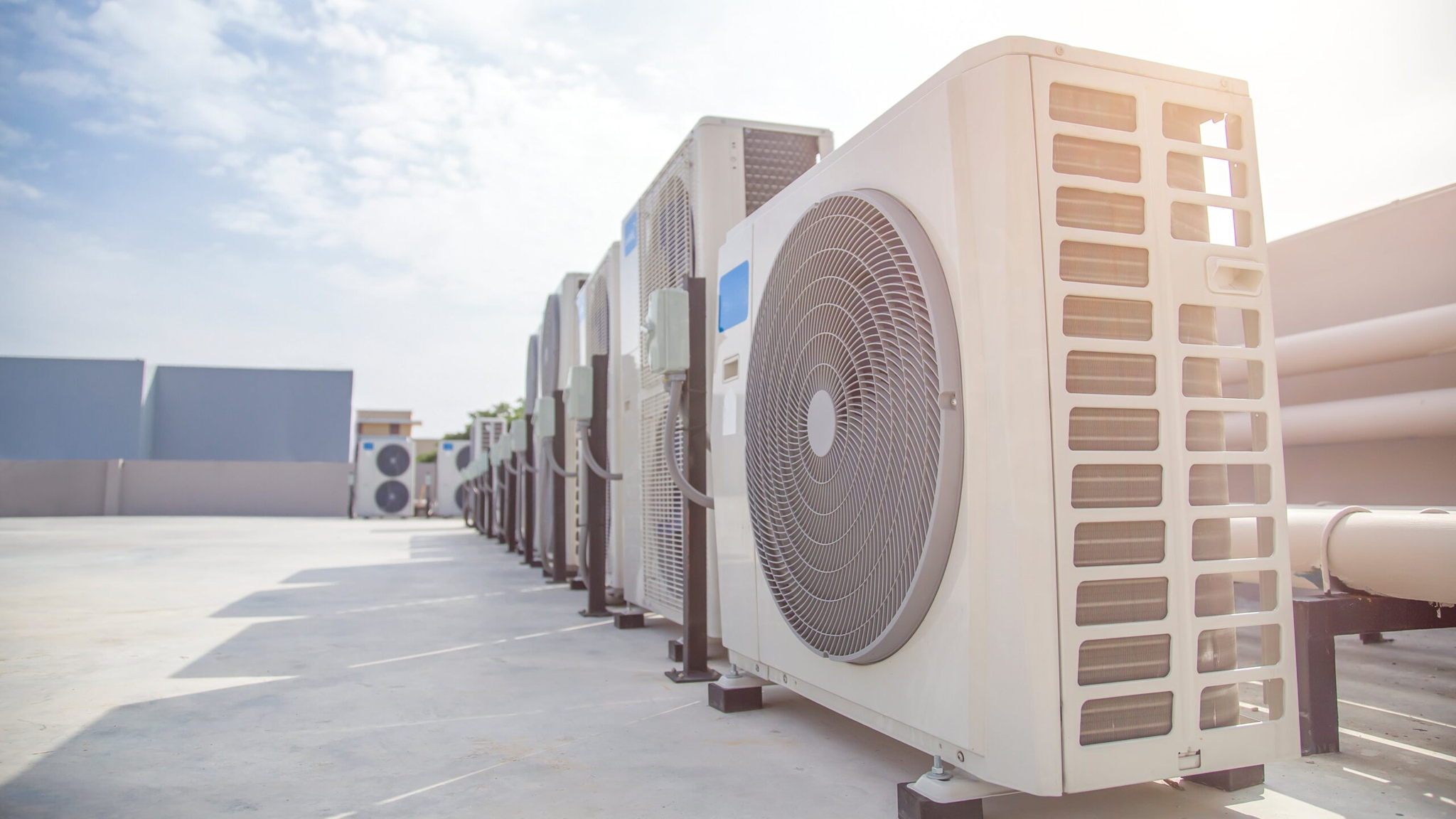 A row of industrial air conditioning units on a rooftop, facing upward, under a clear blue sky. The sun illuminates the units, casting shadows on the flat surface. The perspective emphasizes linearity and repetition.