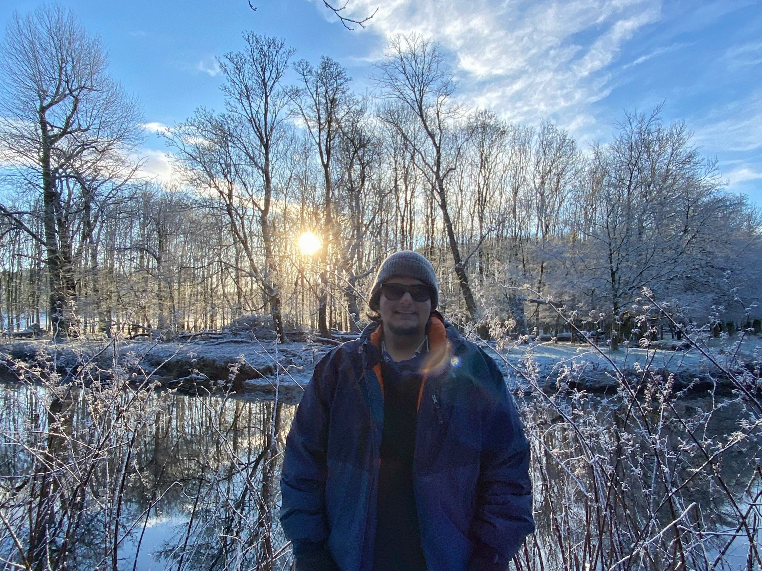 Rishabh stands in the middle smiling with a winter jacket, sunglasses and a hat on. In the background we can see a reflective body of water and the evening sun above his head. We can see trees without leaves covered in snow.