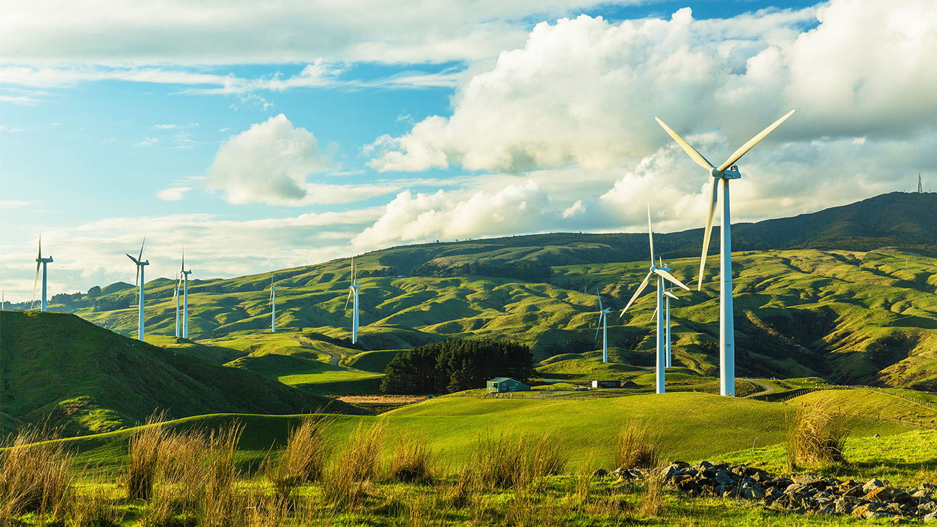 Wind turbines stand on rolling green hills under a partly cloudy sky, generating renewable energy in a rural landscape. The scene is bright and peaceful, with lush grass and distant hills in the background.