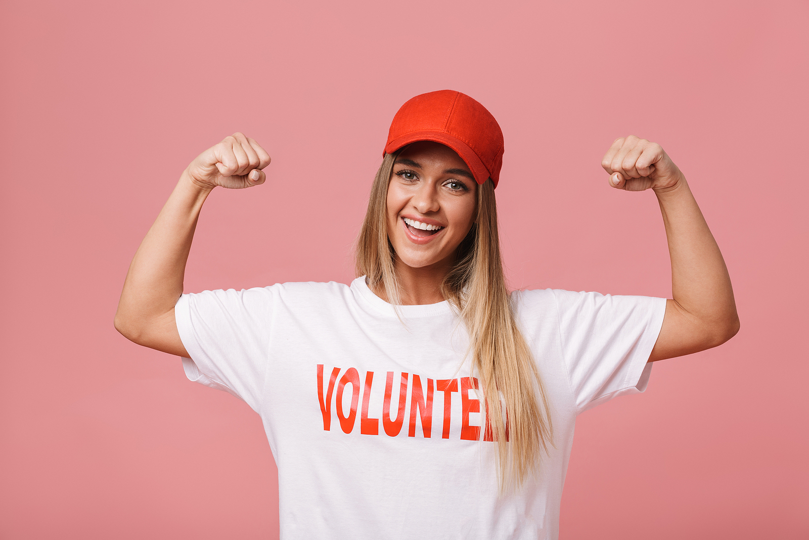 A blond haired woman wearing a shirt that reads volunteer flexes her arms and smiles.