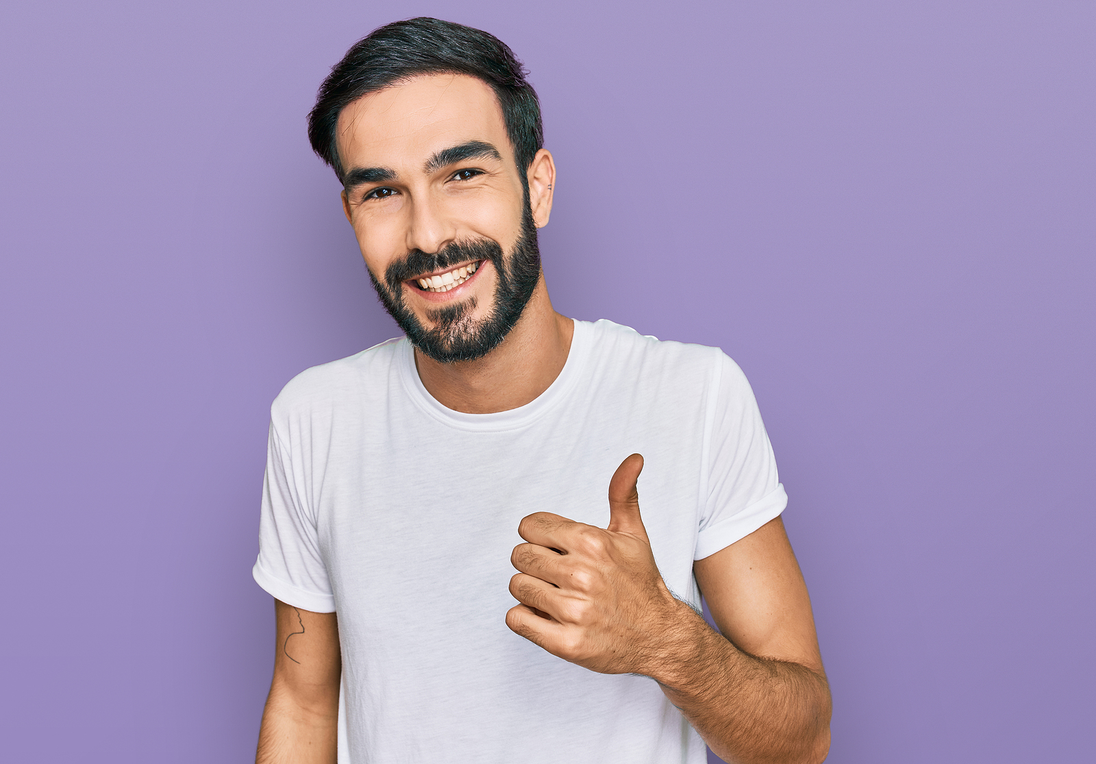 A man smiling with a beard and a white t shirt with thumbs up on purple background.