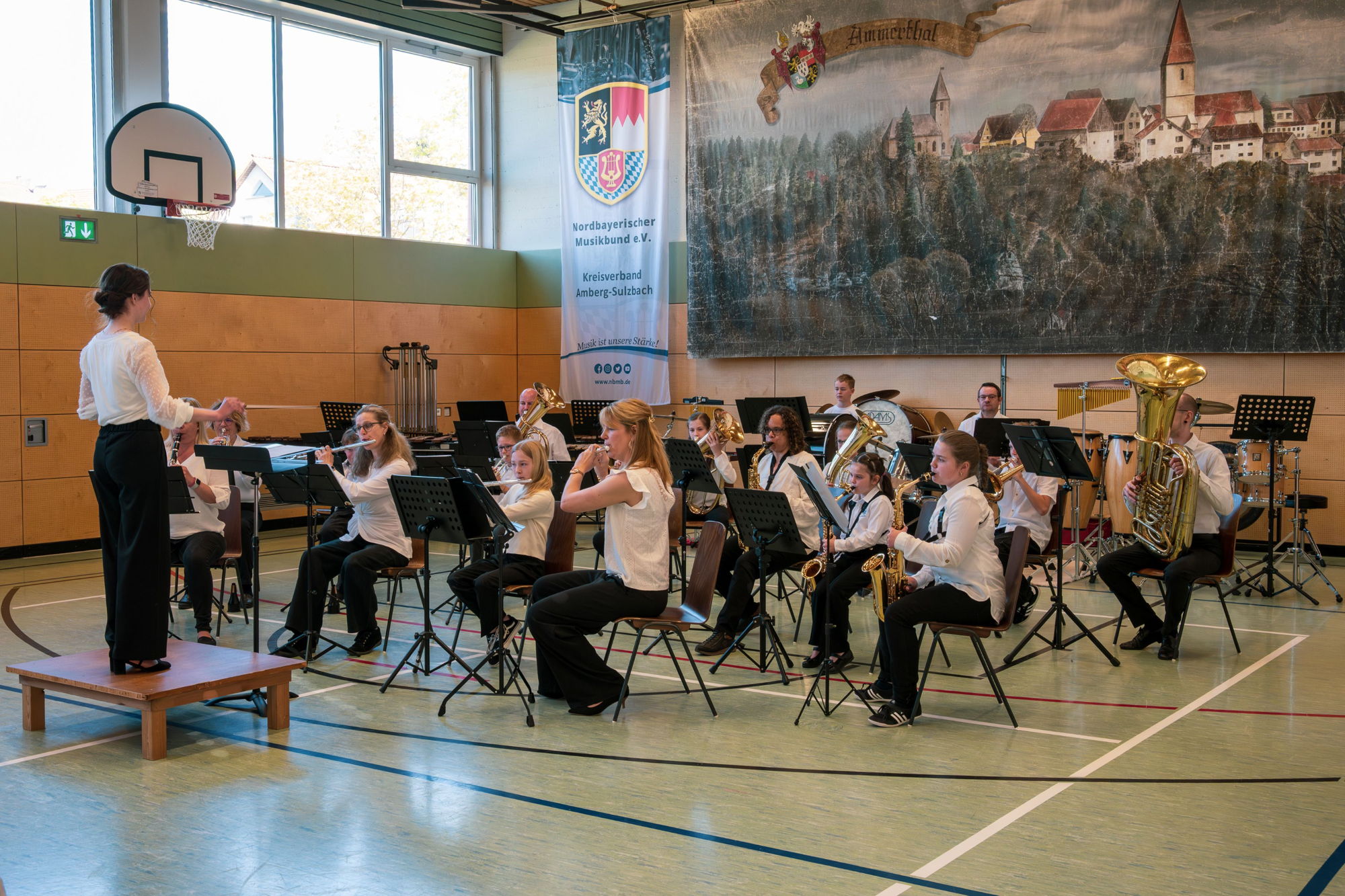 Einige junge Musikerinnen und Musiker in einheitlicher schwarz-weißer Kleidung musizieren in einer Turnhalle. Vor ihnen steht eine junge Dirigentin. 