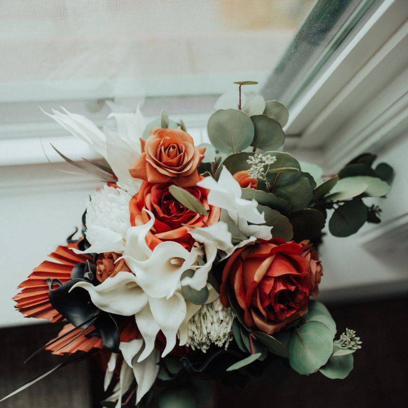 A Halloween themed bouquet rests on a windowsill. It is made of orange roses, white pincushion protea, black and white calla lilies, dried and bleached ferns, and silver coin eucalyptus 