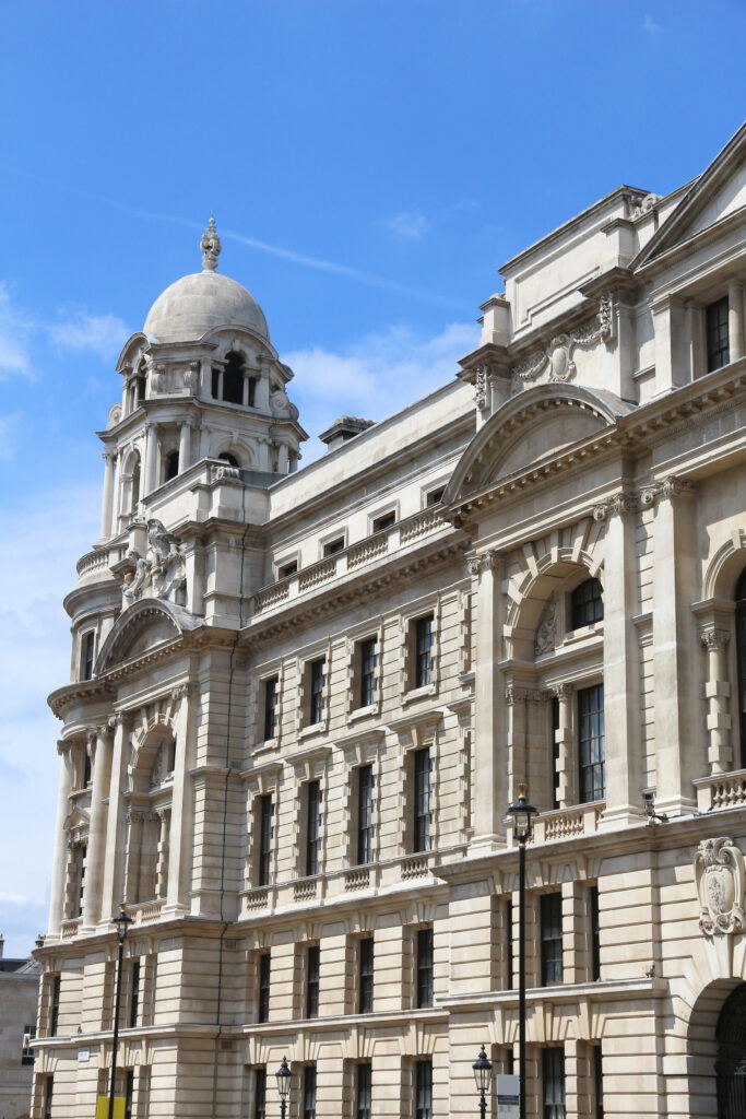 A grand historic building with stone columns, arched windows, and a large dome under a bright blue sky with scattered clouds.
