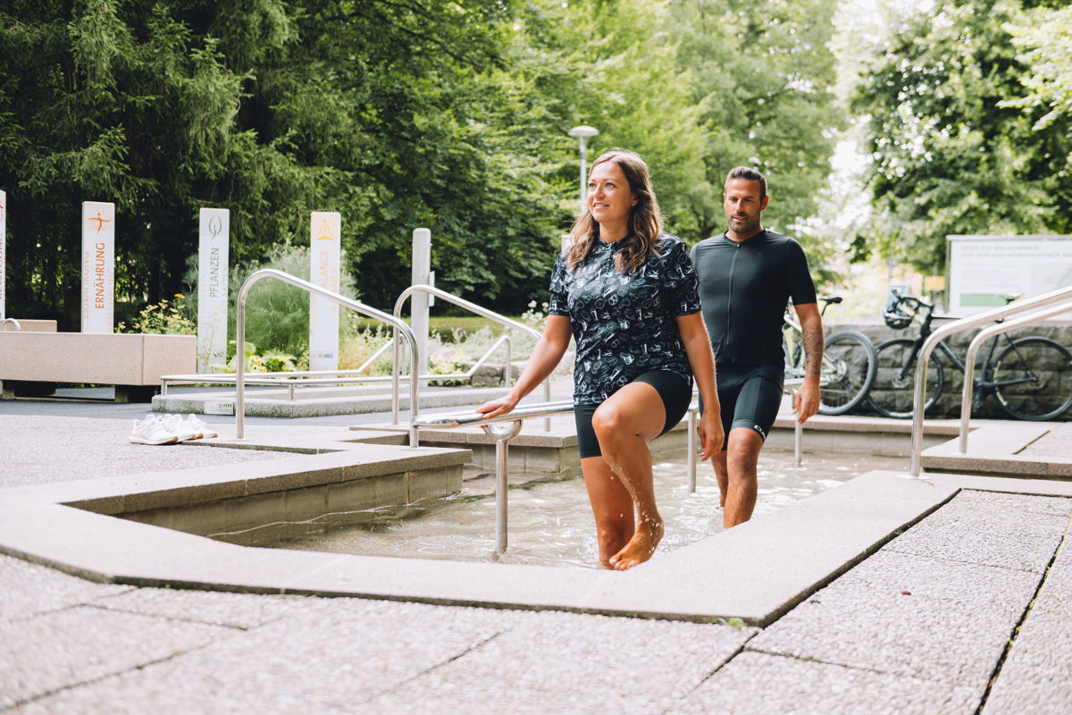 Pärchen im Radoutfit macht eine Pause und nützt diese zum Kneippen im Kurpark.