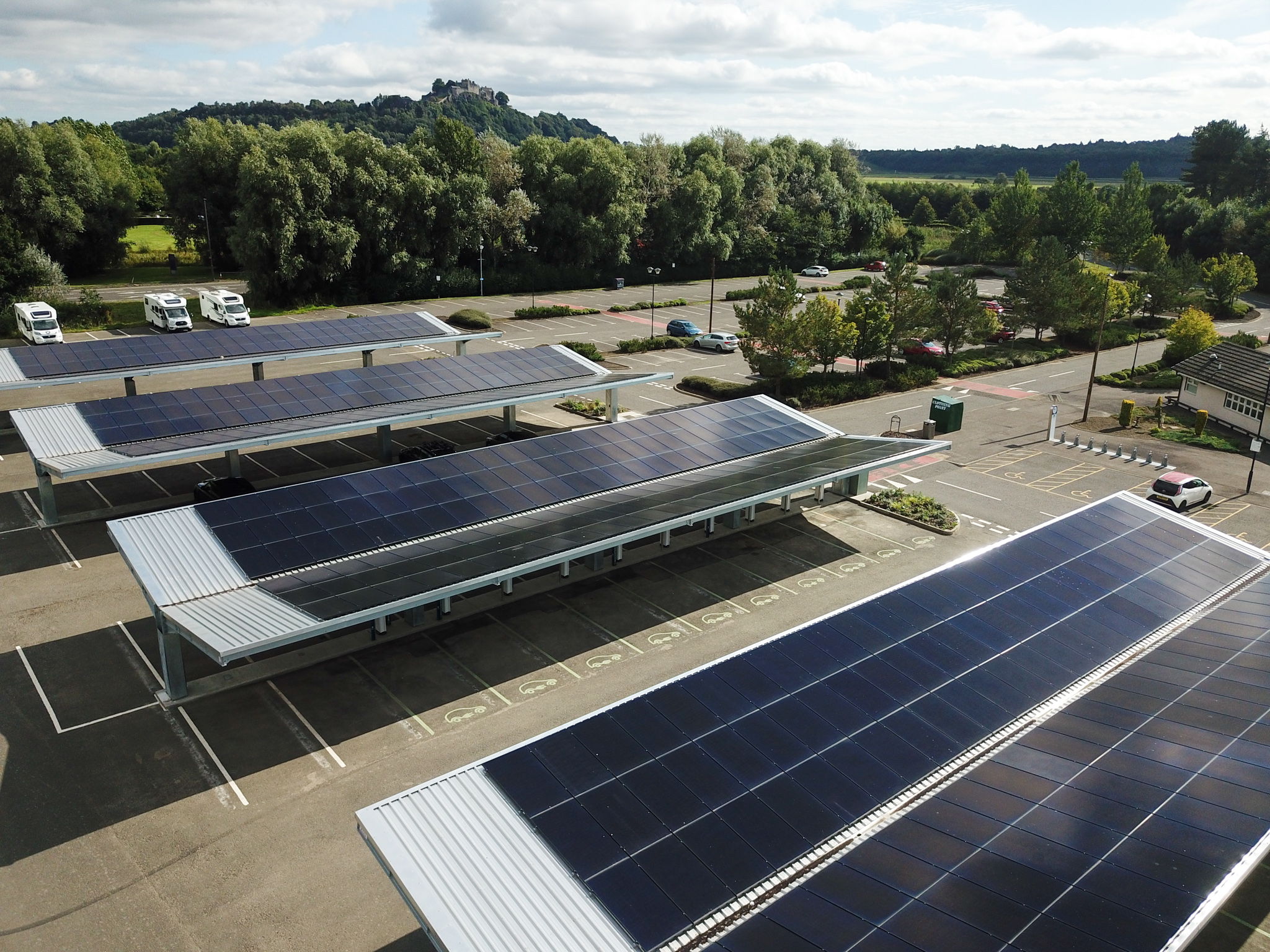 Solar panel canopies cover a large parking lot, with trees, hills, and cars in the background under a partly cloudy sky.