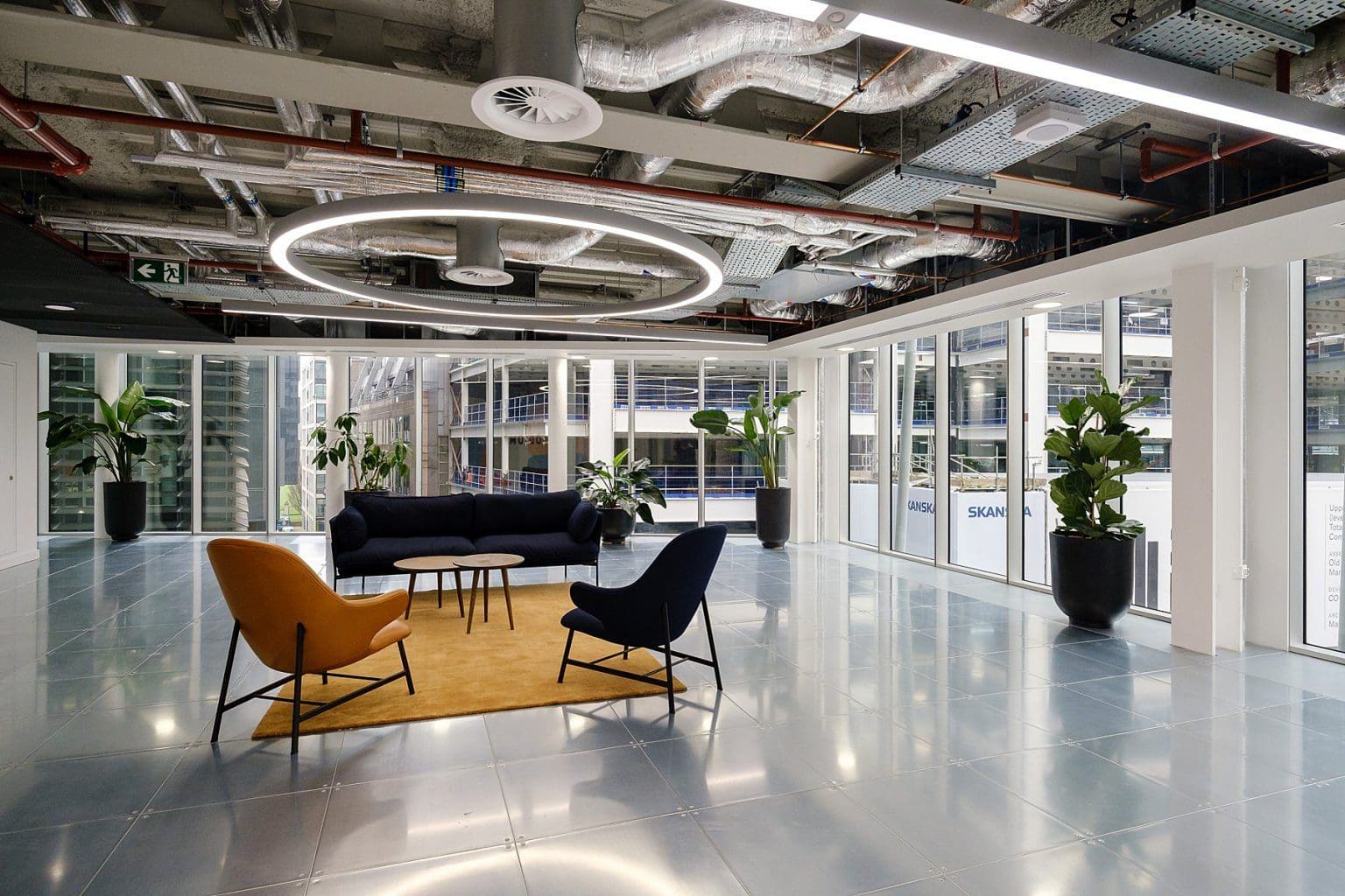 Modern office lounge with large windows, exposed ceiling pipes, a circular LED light, three chairs, a sofa, a small table on a yellow rug, and potted plants, with city buildings visible outside.