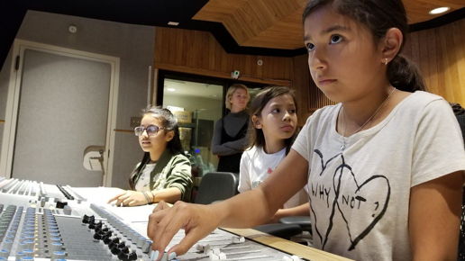 Girls on the Mic — WAM program photo showing three middle-school girls interacting with an audio recording console, with a woman instructor in the background