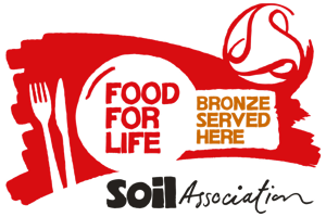 Red and white logo with a fork and knife, the words “FOOD FOR LIFE” in a circle, “BRONZE SERVED HERE” in a gold box, and “Soil Association” in black script below.