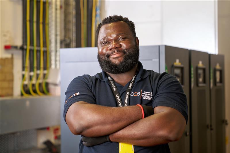 A man with a beard and folded arms smiles confidently in an industrial or technical room, wearing a dark polo shirt and an ID lanyard. Electrical panels and equipment are visible in the background.