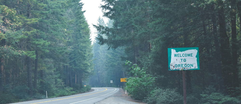a green welcome to oregon sign leading into the forest and paved road