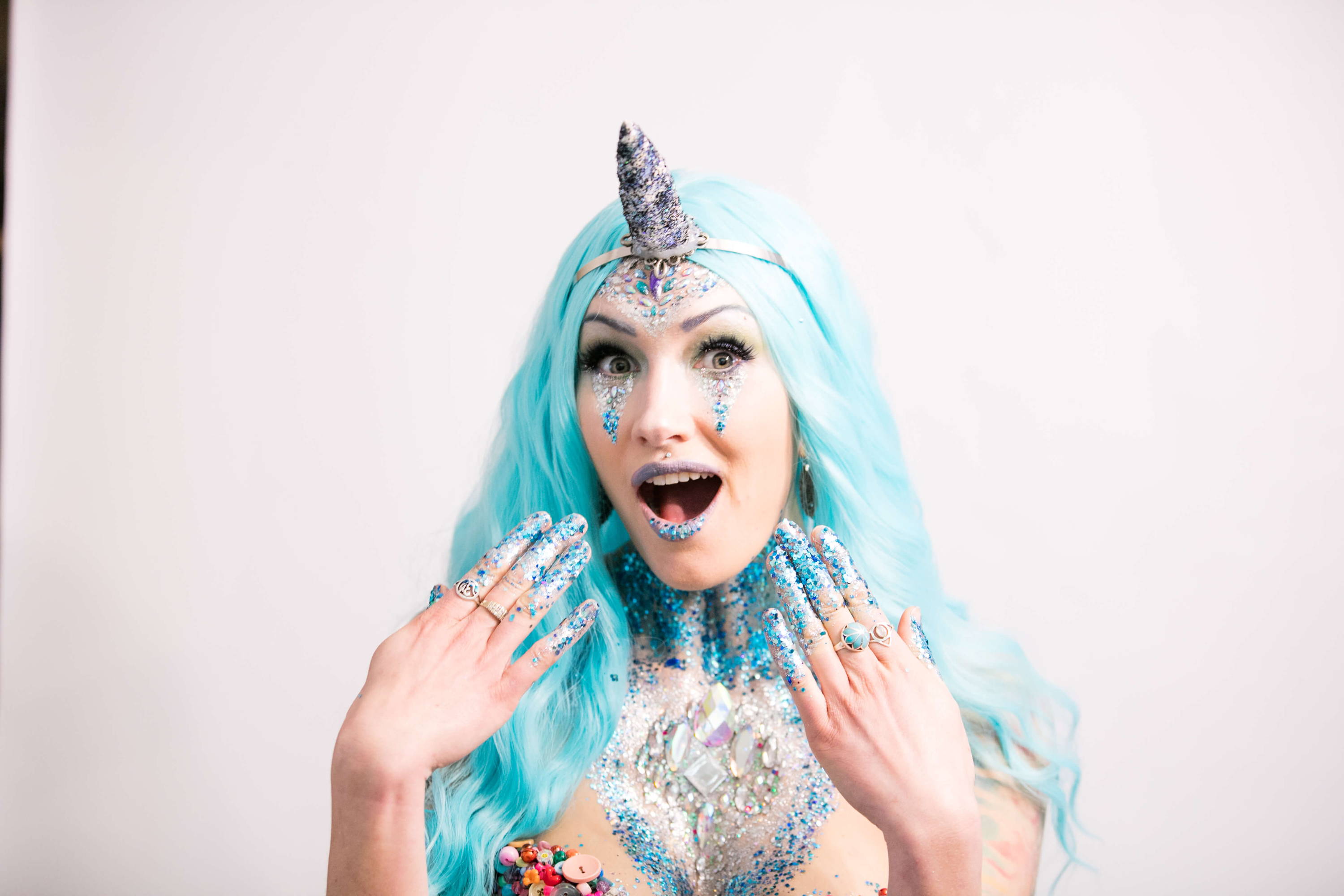 Image shows a women covered in glitter on her face and chest with a unicorn horn on her head. She's holding her hands up in front of her face as if she's surprised and happy.