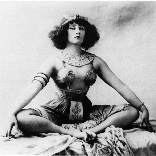 Colette wearing dancers clothes sits with her legs together and her arms on her knees.