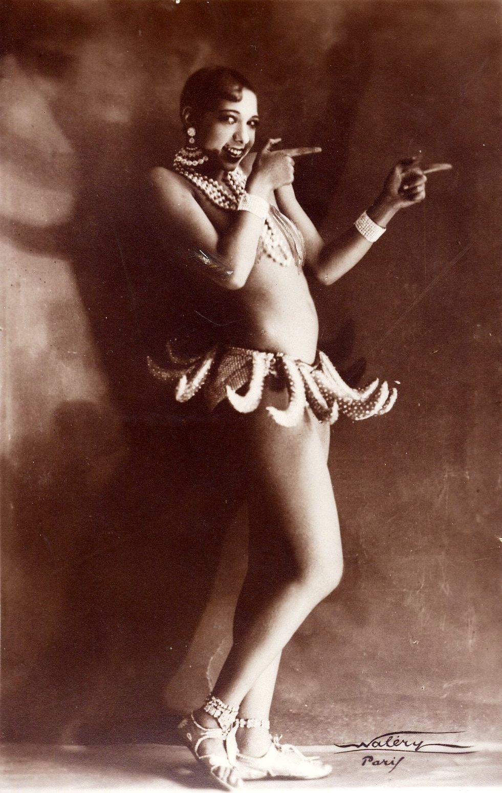 Sepia tone image of Josephine wearing her famous banana dress, with finger guns pointed away as she smiles.