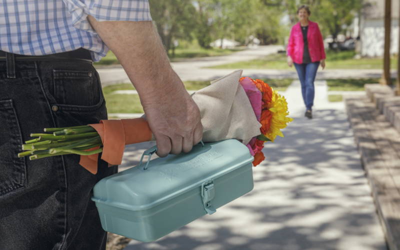 ron holding a bouquet of flowers and a metal toolbox full of quilting tools on his way to surprise Jenny Doan.