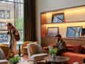 Moderne Hotellobby mit gemütlichen Sitzgelegenheiten, beleuchteten Holzregalen mit Fotos und einem großen Fenster mit Blick auf die Straße.