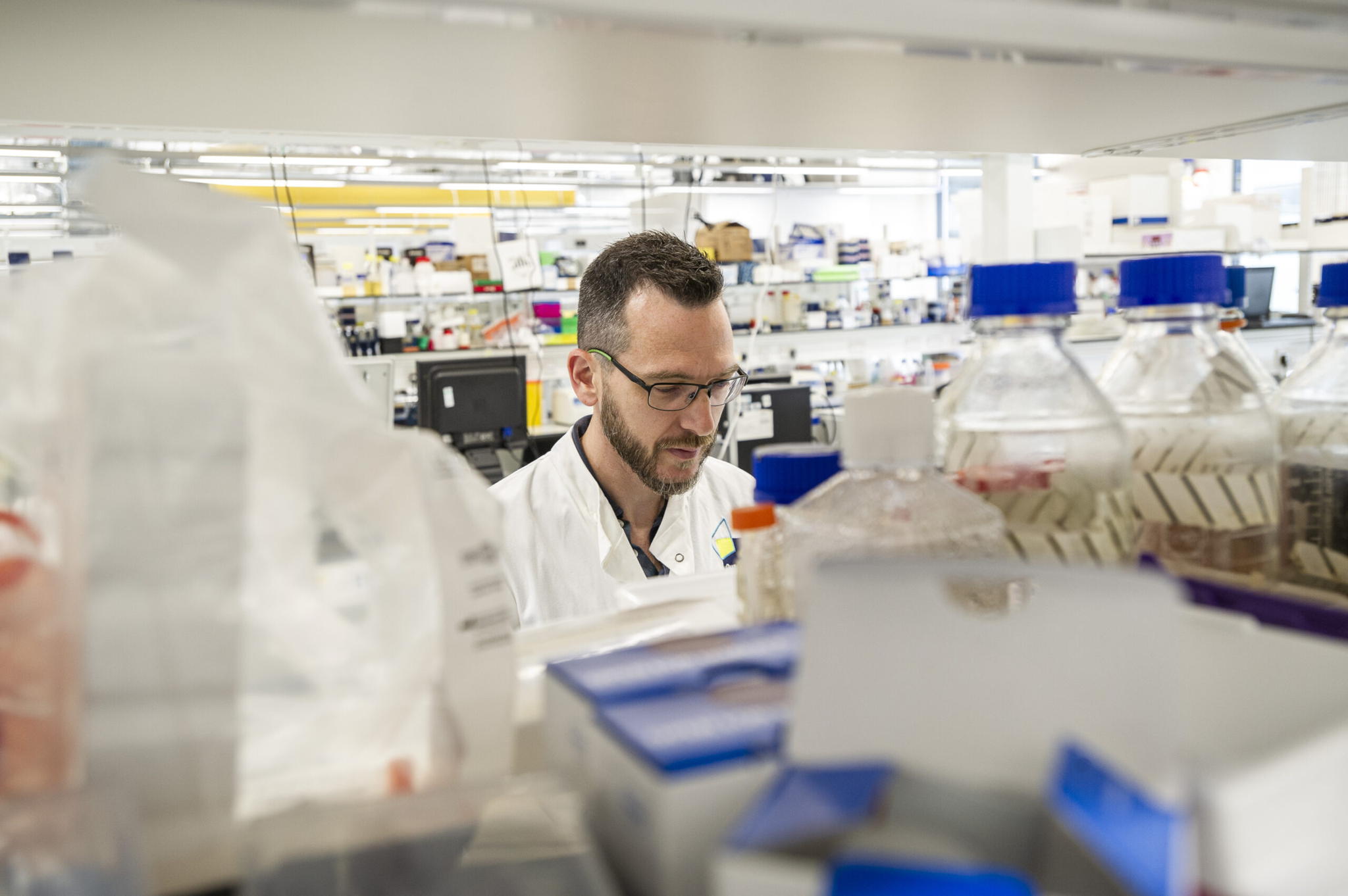 A man in a white lab coat works in a busy laboratory filled with various containers and equipment. He appears focused, surrounded by shelves and counters holding scientific supplies.