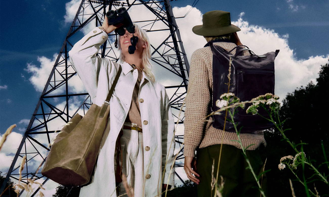 two models wearing ally capellino bags, one is using binoculars and looking into the distance in a white jacket and the other is wearing a beige jumper and looking away from us at a pylon