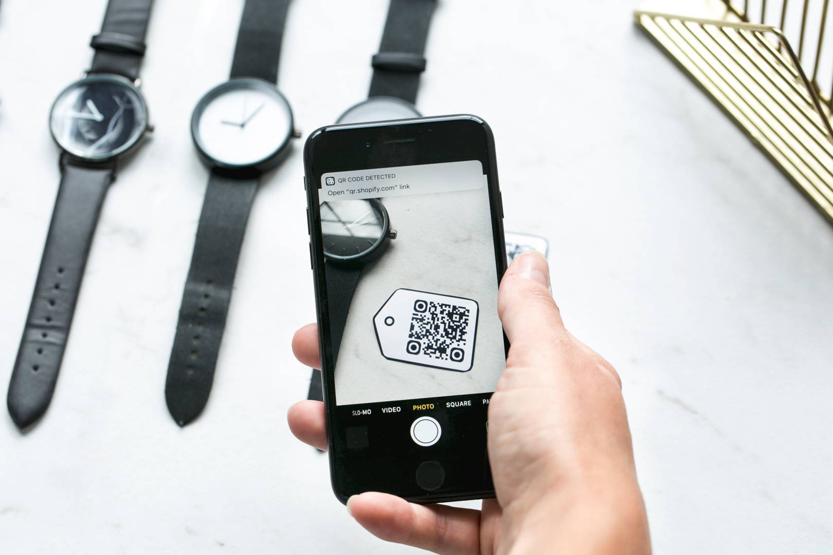 O persoană scanând un cod QR de pe o etichetă cu un telefon mobil, în apropierea a două ceasuri și a unui obiect decorativ auriu.