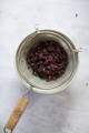 A metal strainer with a wooden handle sits over a bowl, containing strained dark berries on a white surface.