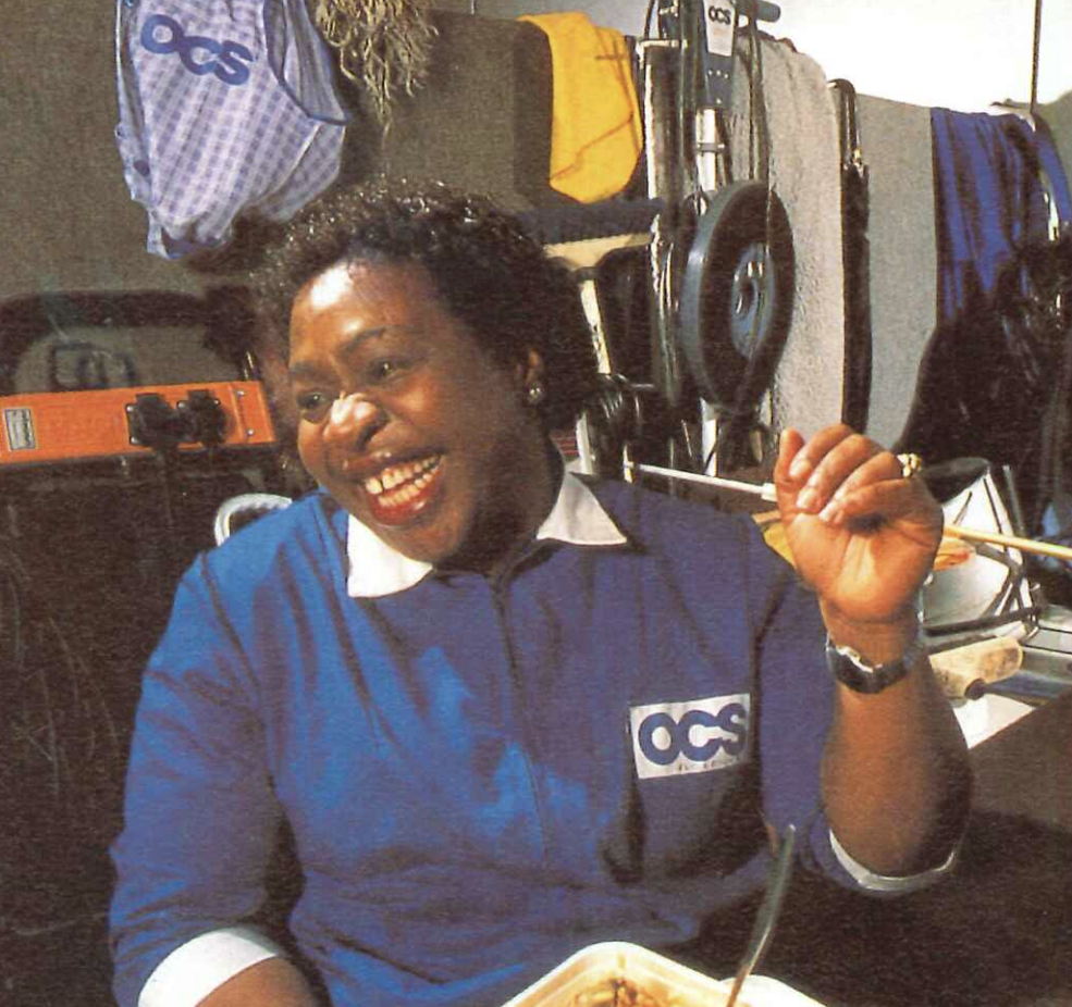 A person wearing a blue uniform with an OCS logo is smiling broadly, seated indoors. They are holding a fork, and various equipment is visible in the background, including wheels and fabric.