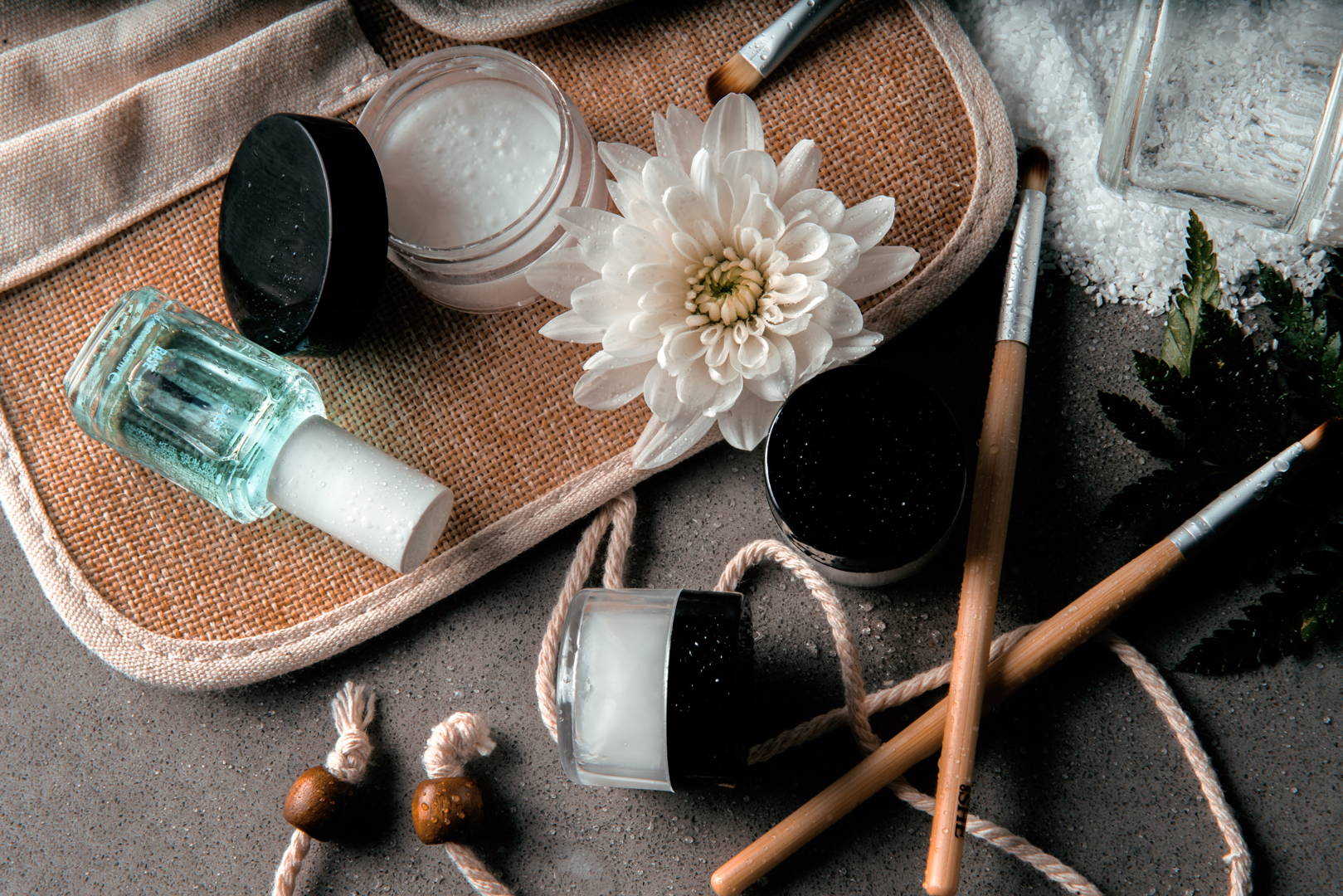 Produse naturale de îngrijire a pielii cu floare de crizantemă albă, borcane din sticlă și pensule cosmetice pe un fundal texturat.