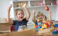 Brother and sister in their room, happily raising their hands in the air while holding wooden toys