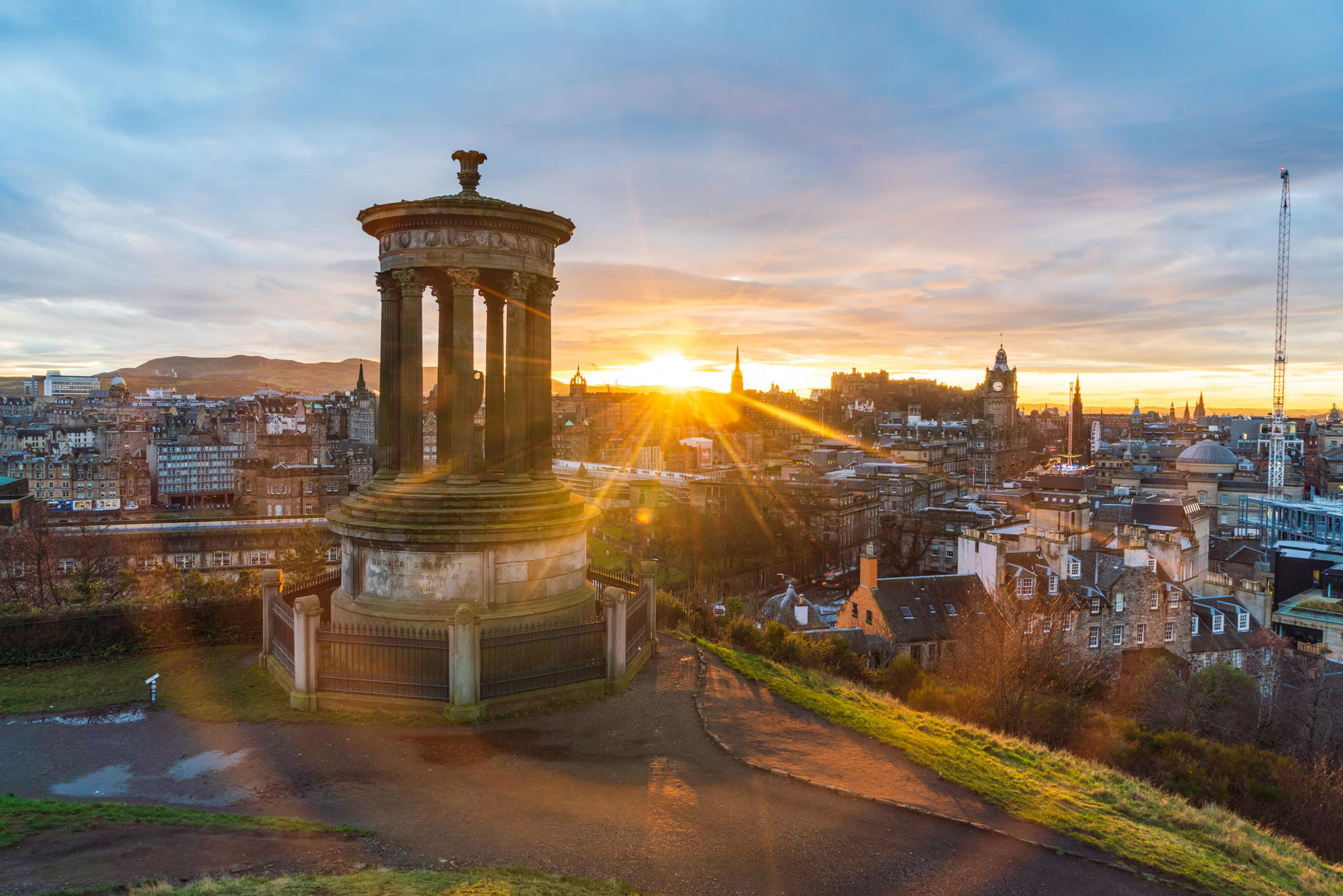 The Dugald Stewart Monument on Calton Hill overlooks Edinburgh at sunrise, with sun rays spreading over the city’s historic buildings and a partly cloudy sky.