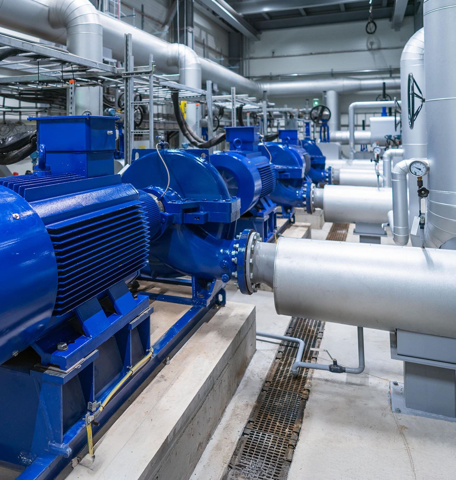 A row of large blue industrial pumps and motors are installed on concrete platforms in a facility, surrounded by metal pipes, gauges, and overhead structural elements.