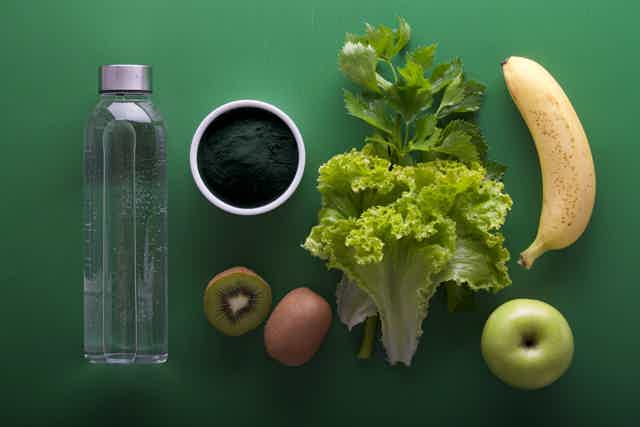 Clear water bottle, spirulina powder, kiwi, lettuce, celery, banana, and green apple arranged on a green surface.