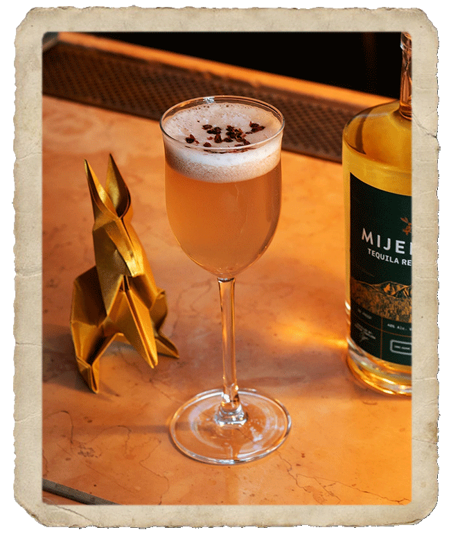 A glass with the prepared Polka Dot cocktail sided by a Mijenta Tequila Reposado bottle and an origami-like rabbit miniature