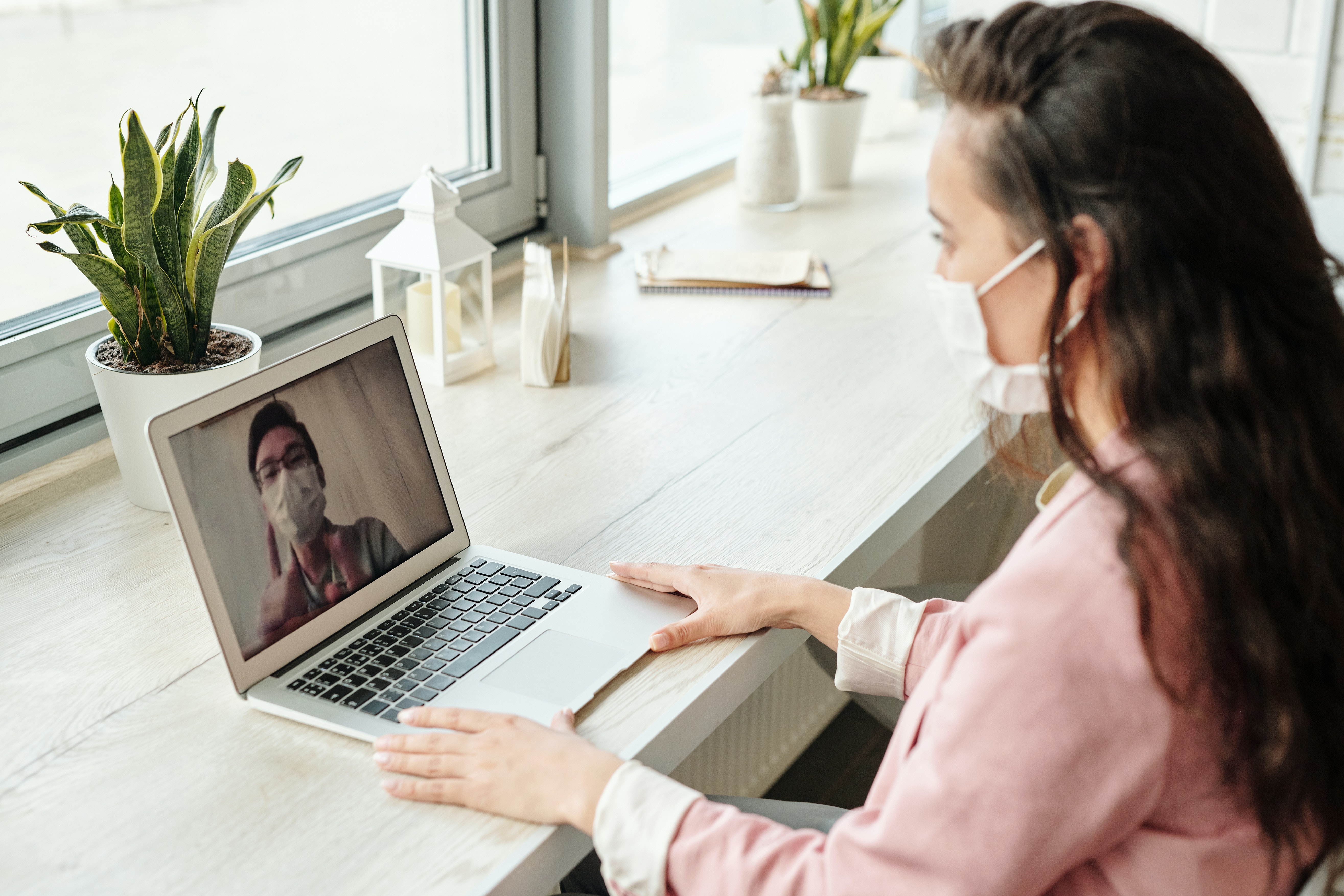 Image of a woman wearing a facemask and sitting on a desk, with her laptop on a video call with someone else with a facemask.