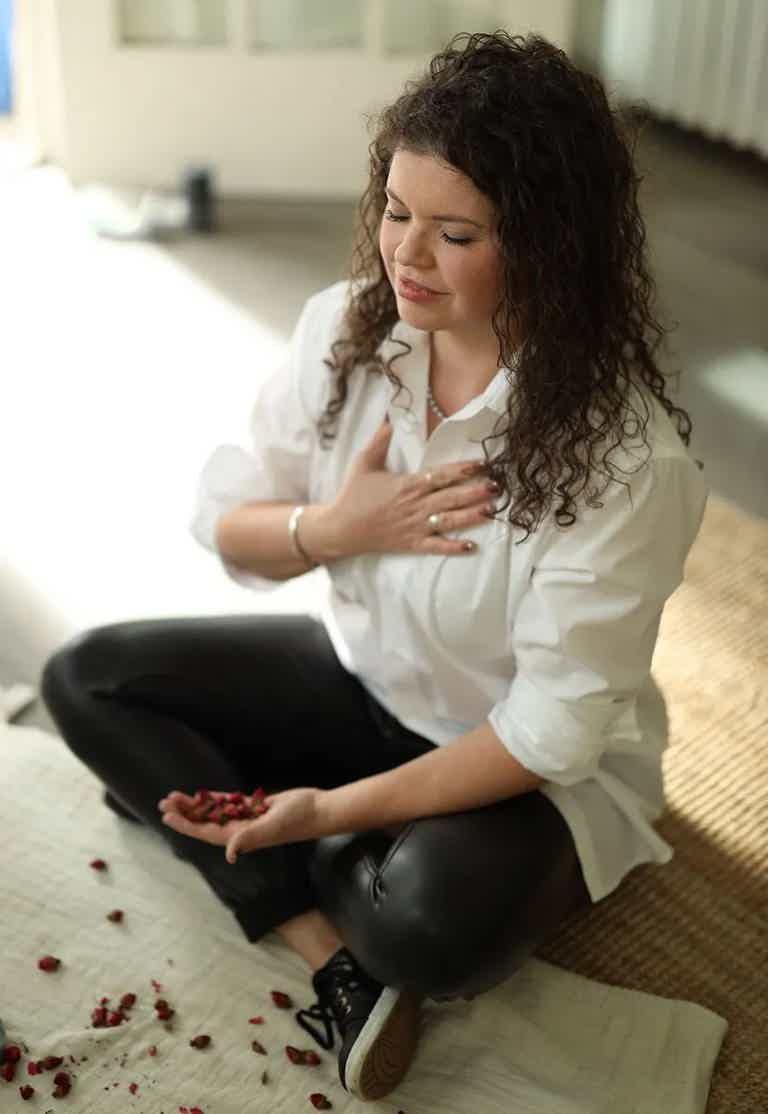 Serene woman sitting cross-legged on rug, hand over chest, petals scattered, soft natural light.