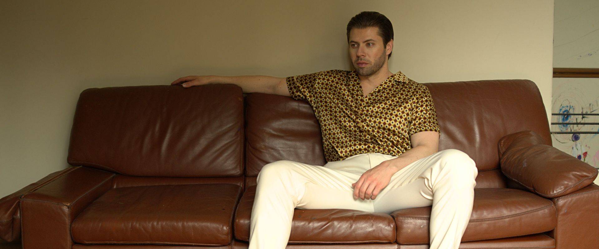 all collections product page header image of a man wearing a silk shirt and white pants sitting on a sofa