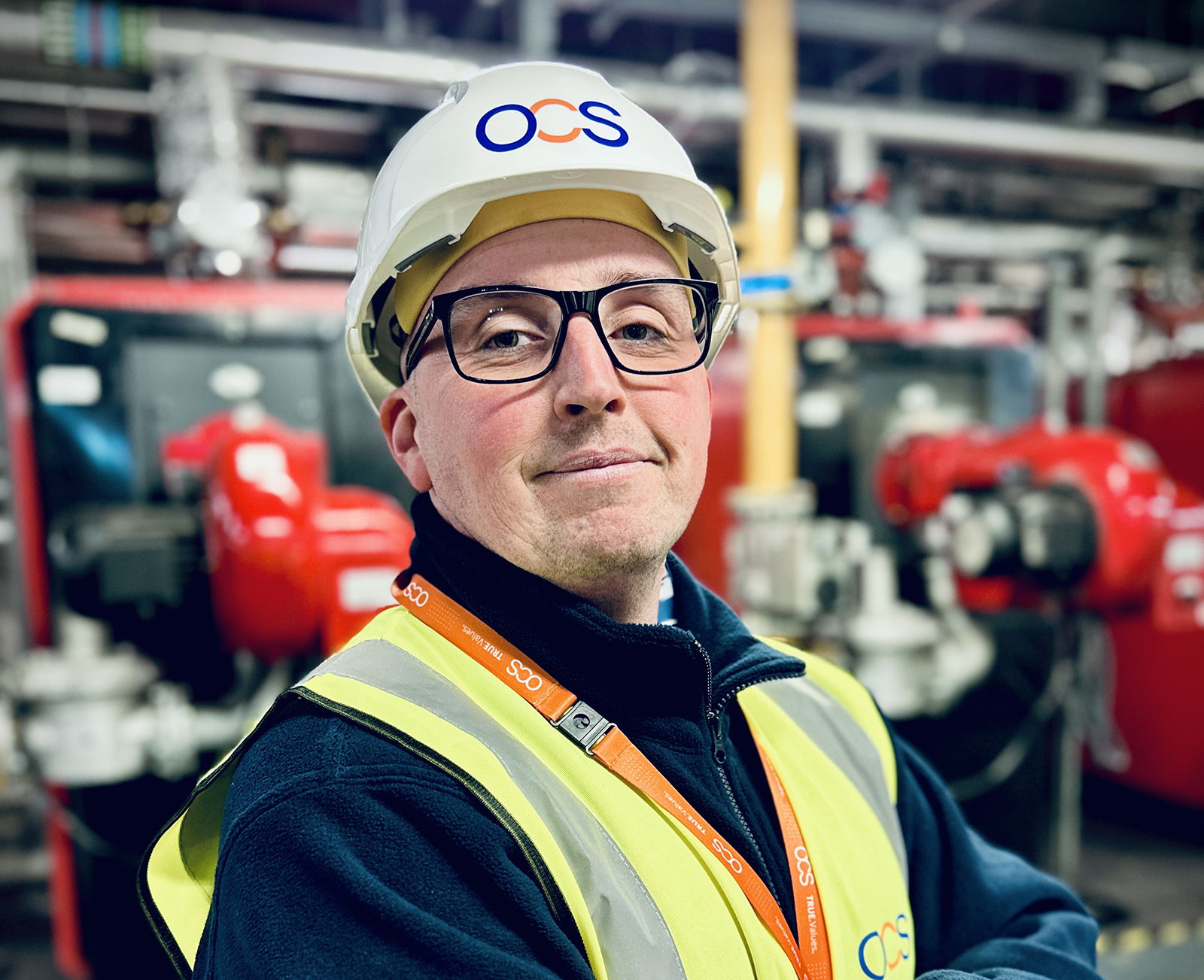 A man wearing a white hard hat, safety glasses, a high-visibility vest, and an OCS lanyard stands confidently with arms crossed in an industrial setting with red machinery in the background.