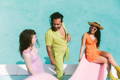 a woman wearing a straw hat and mustard boots sits on a pink slide wearing sunglasses while a man in a chartreuse jumpsuit speaks with a curly haired woman wearing a lilac jumpsuit for "poolside pride: a slim aarons-inspired photo campaign."