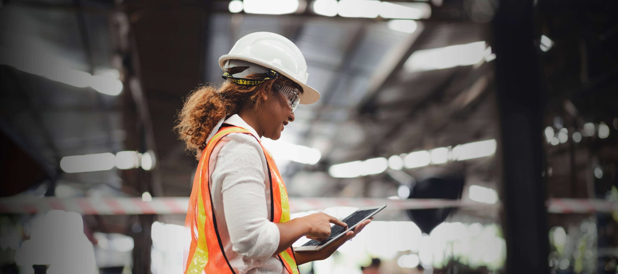 An OCS colleague in a hard hat and high-visibility vest holds a tablet inside a warehouse, showcasing expertise in distribution and logistics, as well as fabric and building maintenance and refurbishment.