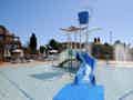 Ein flaches Kinderbecken mit blauer Wasserrutsche, Kippeimer und Wasserspielen, umgeben von Sonnenliegen unter blauem Himmel.