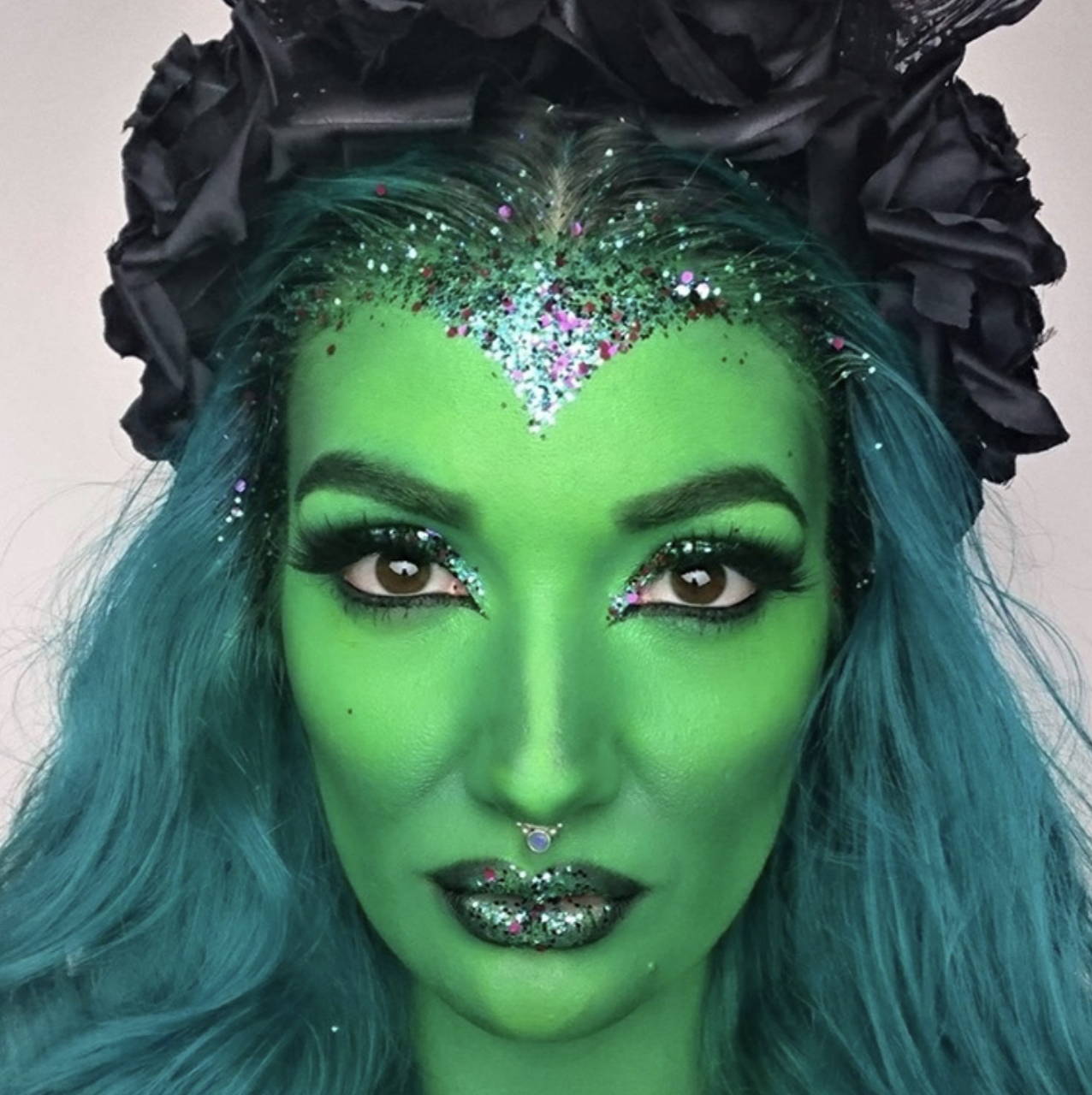A green halloween makeup look inspired by Elphaba from wicked. Model has green face paint topped off with green and purple bioglitter.