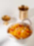 A white bowl with handles containing bright orange Indian sweet garnished with cashews and dried fruit, with a brass lamp and cup in the background.