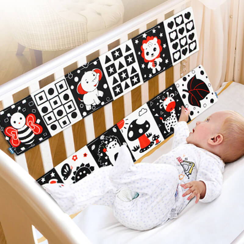 Un nouveau-né dans un berceau, regardant des cartes noires et blanches à contraste élevé d'un livre de contraste pour nouveau-né Montessori InvenToy comportant des illustrations d'animaux, attachées à la balustrade du berceau.
