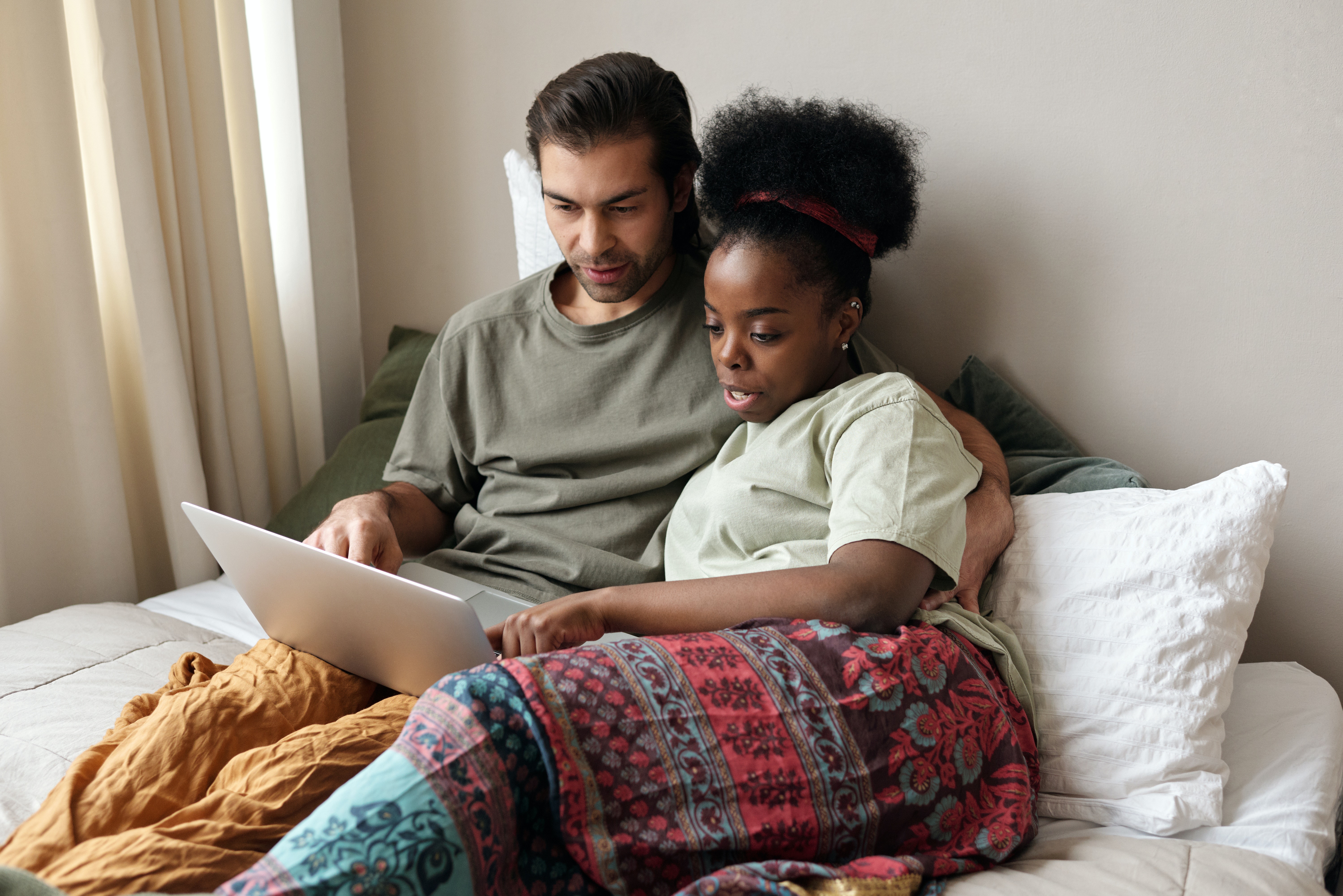 A multi racial couple lays in bed together looking at a laptop and talking about something they see.