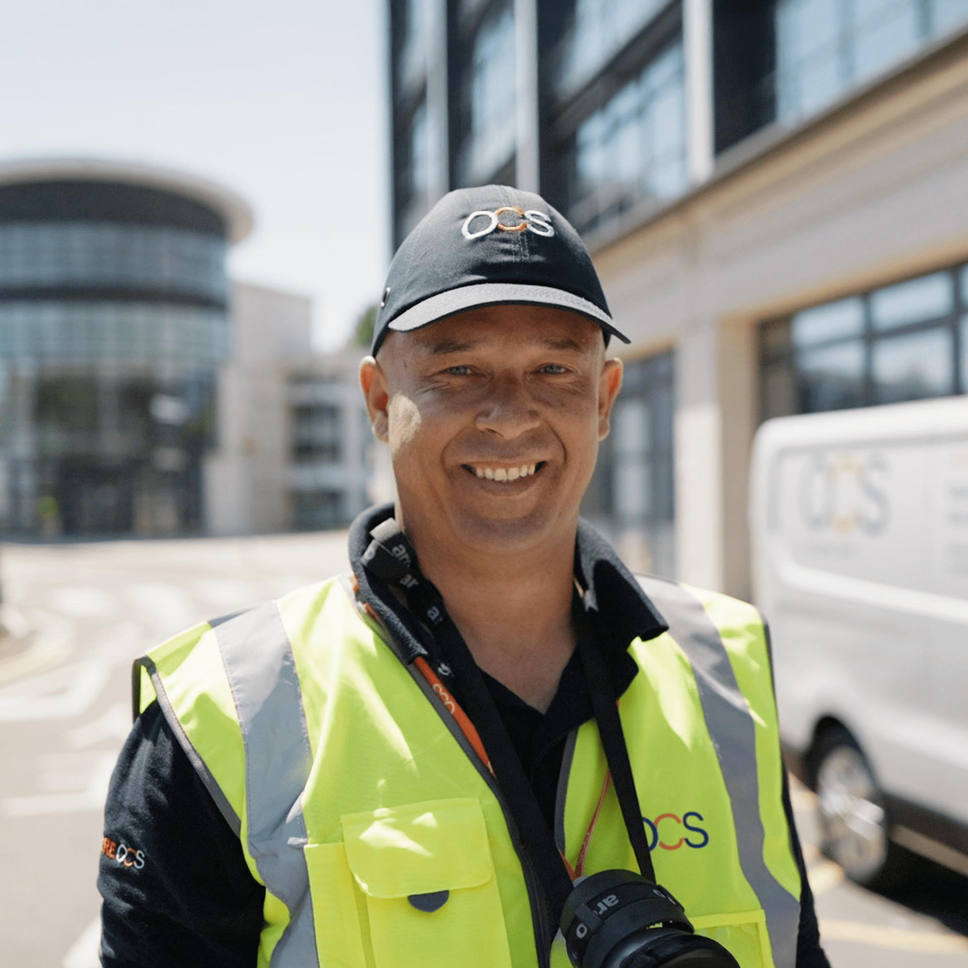 A man wearing a yellow safety vest and a black cap with OCS on it smiles at the camera, standing outdoors near a modern building and a white van with the OCS logo in the background.