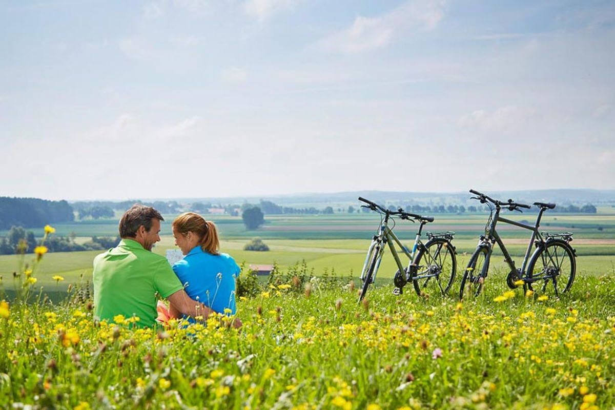 Ein Pärchen sitzt auf einer Wiese mit gelben Blumen, rechts neben ihnen stehen zwei Fahrräder. Im Hintergrund sieht man Hügel und Bäume.