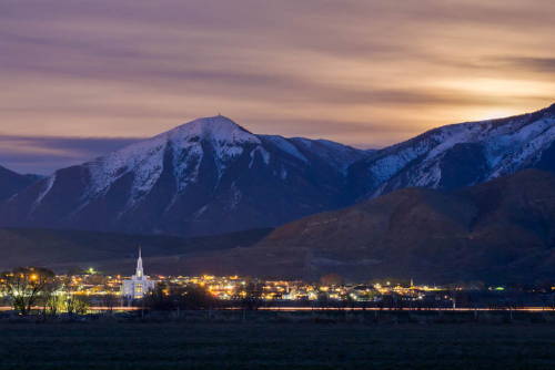 Distant photo of the Payson Utah Temple glowing amid city lights beneath the mountains